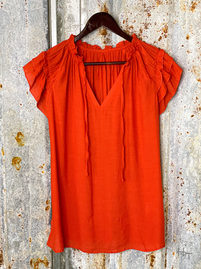 Photo of the front view of a terra cotta red short sleeve top with ruffle accents on the sleeves and a V neck on a hanger with a tin background