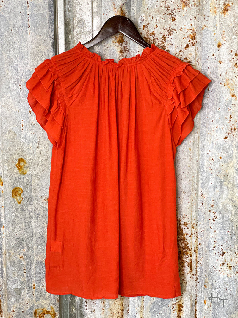 Photo of the view of a terra cotta red short sleeve top with ruffle accents on the sleeves on a hanger with a tin background