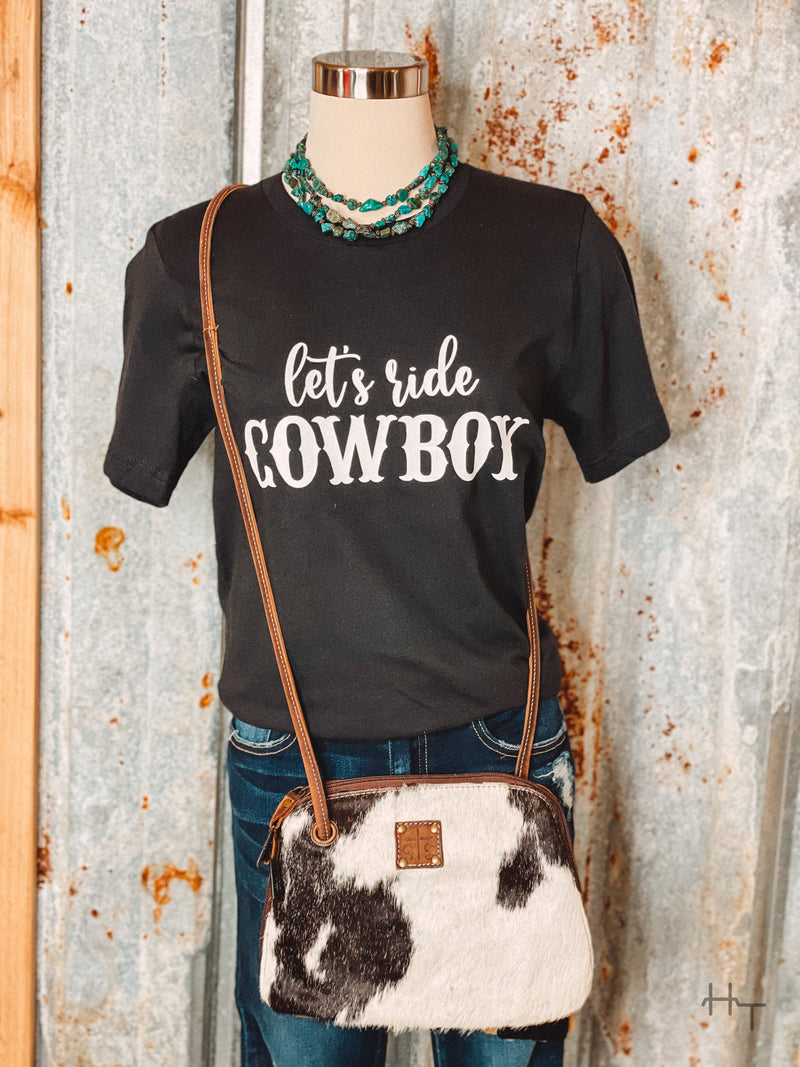 Photo of black tee shirt with Let's Ride Cowboy on front in white paired with turquoise necklace and black and white cowhide purse on a mannequin with tin background