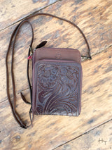 Photo of front of chocolate brown leather purse with tooled leather panel on front pocket with long crossbody strap with wood background