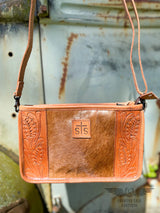 Close up image of the front of the Yipee Kiyay Claire crossbody purse.  The purse has a brown cowhide panel in the middle with floral tooled veggie tan leather on each side and a leather patch in the middle with the STS brand.