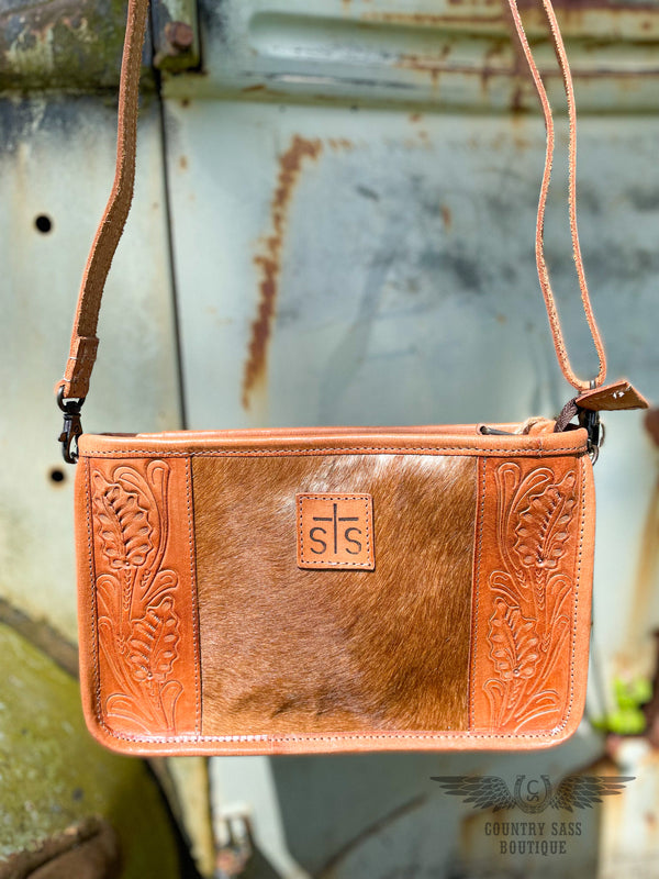 Close up image of the front of the Yipee Kiyay Claire crossbody purse.  The purse has a brown cowhide panel in the middle with floral tooled veggie tan leather on each side and a leather patch in the middle with the STS brand.